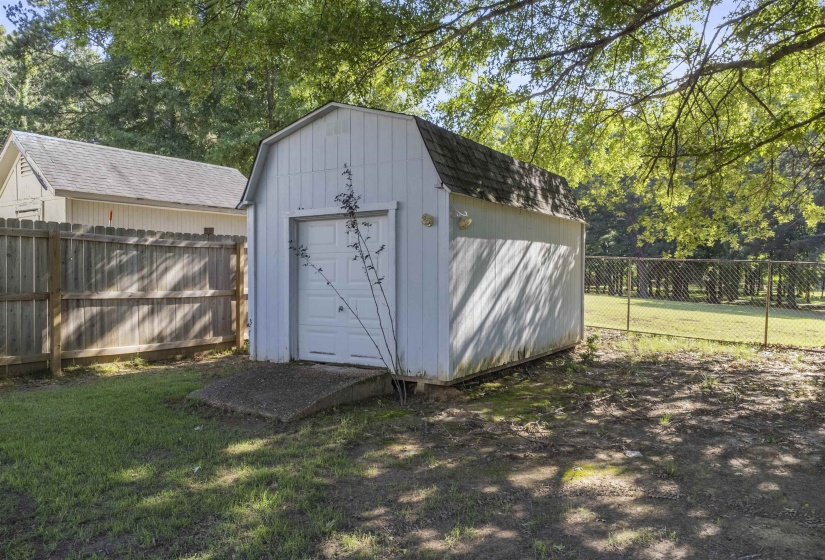 White utility shed with a gambrel roof and a rollup garage door, featuring a concrete ramp entry