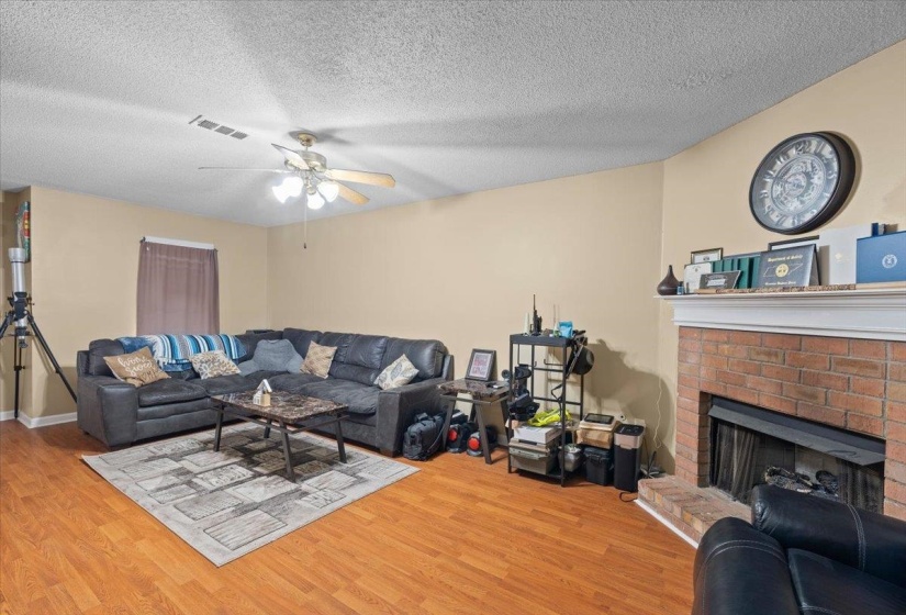 Living space featuring wood-finish flooring, a ceiling fan with light fixture, and a brick-faced fireplace with a white mantel