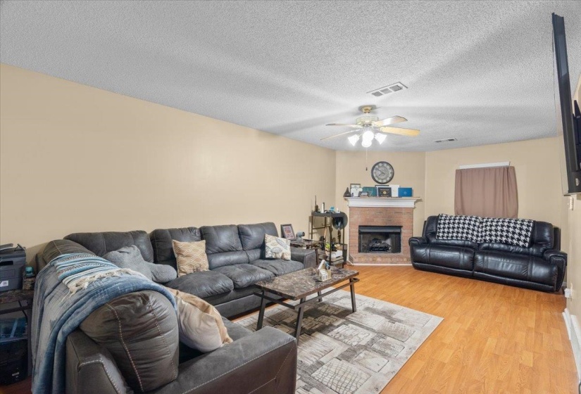 Living area featuring wood-finish flooring, a ceiling fan with integrated lighting, and a brick fireplace with a white mantel