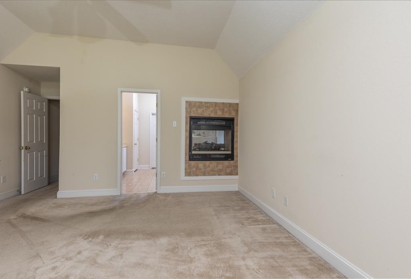 Spacious interior room featuring a wall-mounted fireplace with a tiled surround, vaulted ceilings, light-toned carpeting, and a white baseboard trim