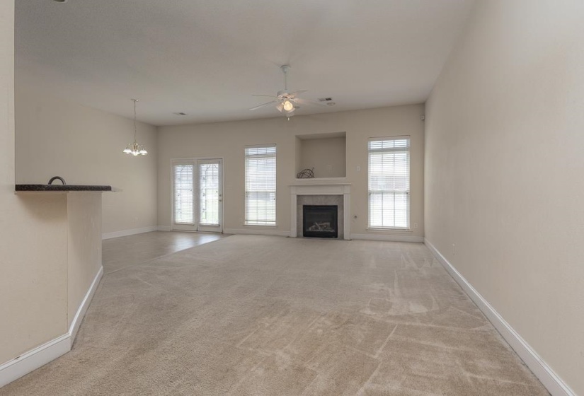 Spacious carpeted room featuring a built-in fireplace with a mantel, multiple windows with blinds, double glass doors, and a ceiling fan