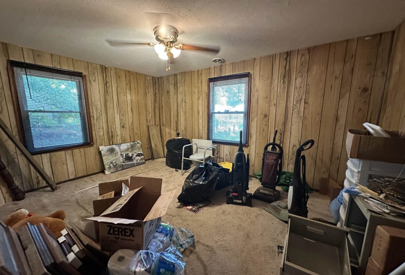 Room featuring vertical wood paneling, two windows with blinds, a ceiling fan with light fixture, and a textured ceiling