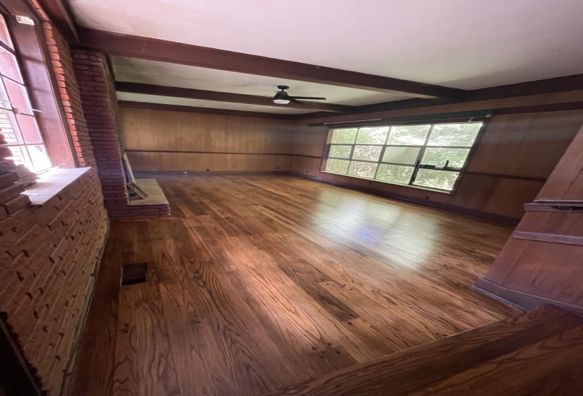 Spacious room featuring hardwood flooring, exposed ceiling beams, a brick fireplace surround, wood paneling, and expansive windows