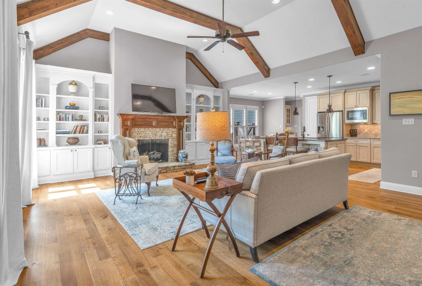 Spacious living area featuring exposed ceiling beams, a stone-surround fireplace with wood mantel, built-in cabinetry, wood-finish flooring, and a ceiling fan
