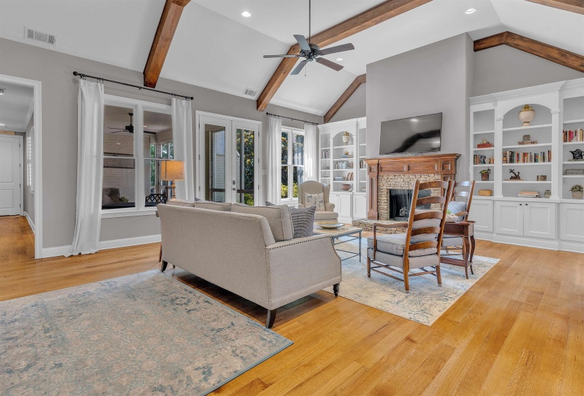 Hardwood flooring throughout a spacious living area featuring vaulted ceilings with exposed wood beams, a brick fireplace with a wood mantel, and built-in shelving with cabinetry