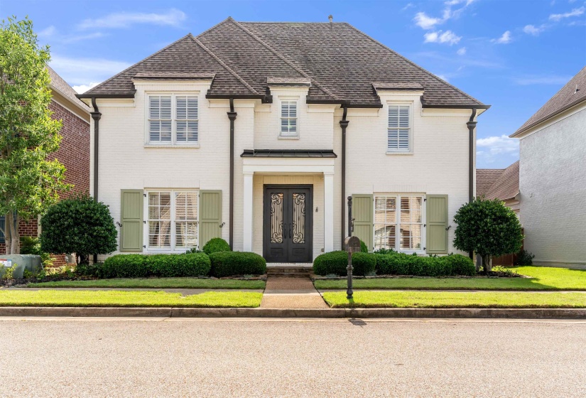 White brick exterior featuring a dark gabled roof, prominent dormer windows, decorative black downspouts, and light green shutters