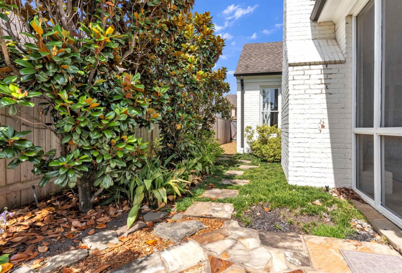 Side yard featuring a stone pathway, mature plantings, white brick exterior, a window with white trim, and a shingled roofline