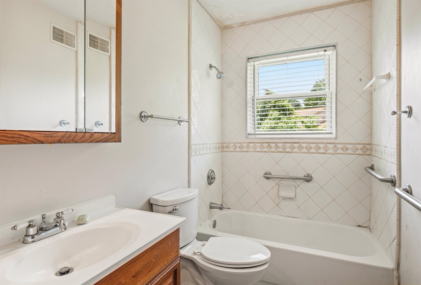 Bathroom featuring a window with blinds, white tile shower surround with decorative accent, built-in bathtub, and a vanity with a white countertop and wood-finish cabinetry