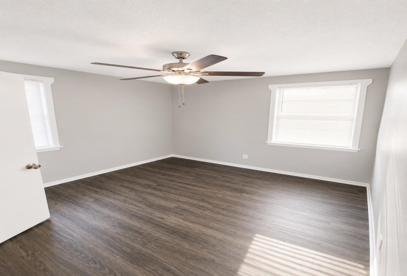 Spacious room featuring wood-finish flooring, two windows with white blinds, a ceiling fan with integrated lighting, and neutral wall paint