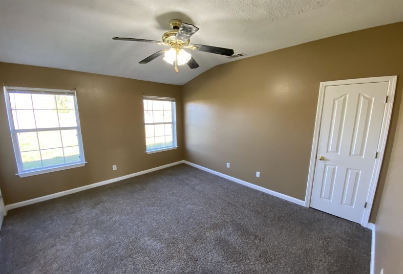 Room featuring vaulted ceiling, ceiling fan with light fixture, two windows with white trim, tan painted walls, and textured carpet flooring