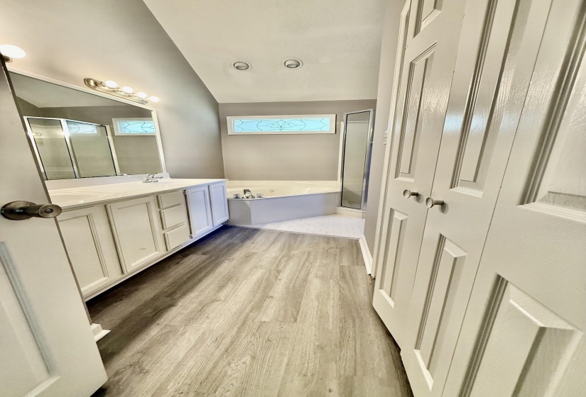 Bathroom featuring wood-finish flooring, a double vanity with white cabinetry, a corner soaking tub, and a separate glass-enclosed shower