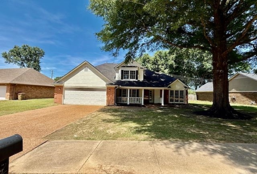 Brick and siding exterior featuring a covered front porch with white railings, an attached garage, a gravel driveway, and a large mature tree