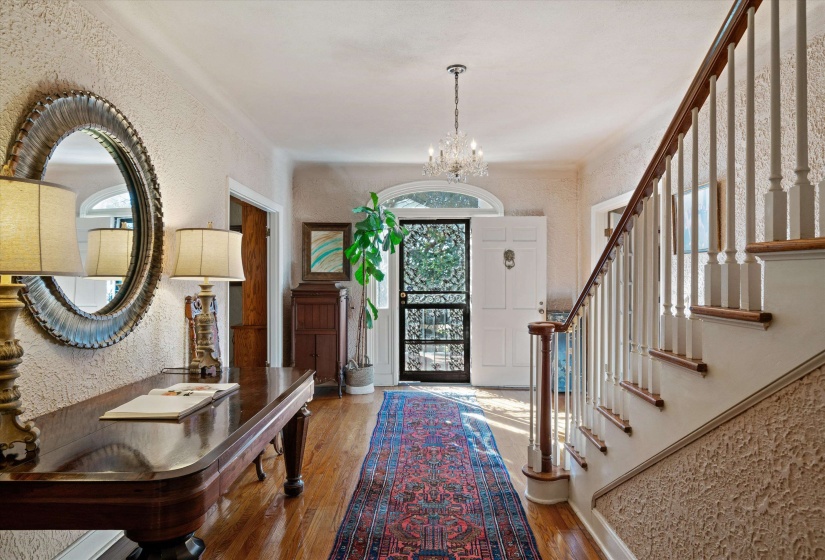 Welcoming entryway featuring hardwood flooring, a detailed staircase with wood handrail, an arched transom window above the front door, textured wall finishes, and a crystal chandelier