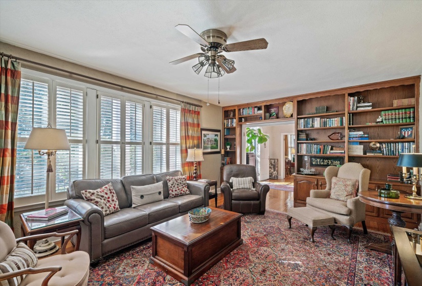 Living area featuring a full wall of built-in wood shelving, multiple panel windows with white plantation shutters, and light wood-finish flooring visible in the background