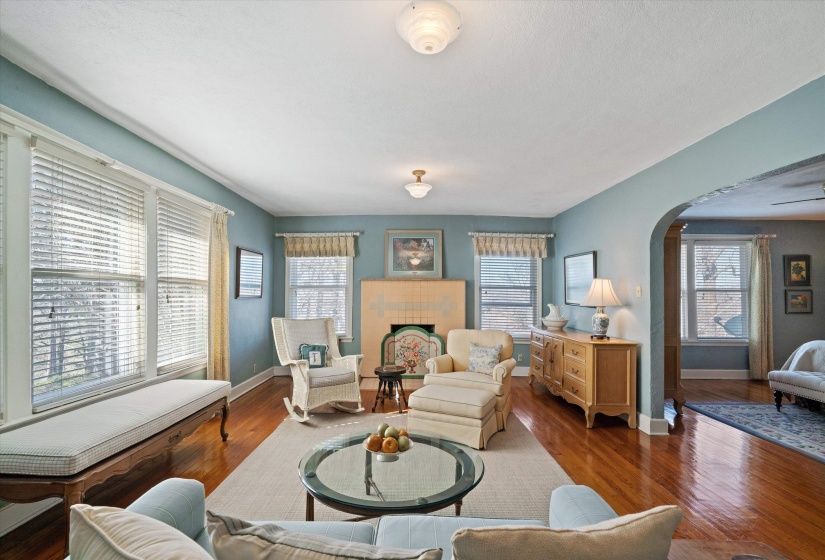 Hardwood flooring throughout the room, multiple windows with blinds, built-in bench seating, an arched doorway, and a tiled fireplace