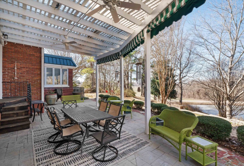 Expansive paved patio featuring a white pergola with two ceiling fans, a striped retractable awning, and mature landscaping