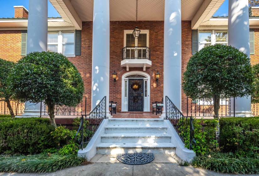 Brick exterior featuring a portico with large white columns, a staircase with black wrought iron railings, a dark entry door with decorative ironwork, and a second-story balcony