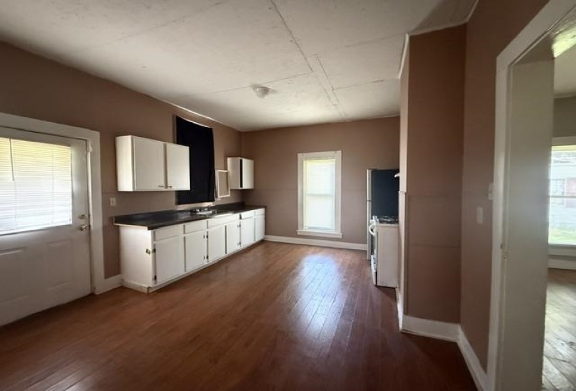 Kitchen area featuring wood-finish flooring, white cabinetry, dark countertops, and a white exterior door with horizontal blinds