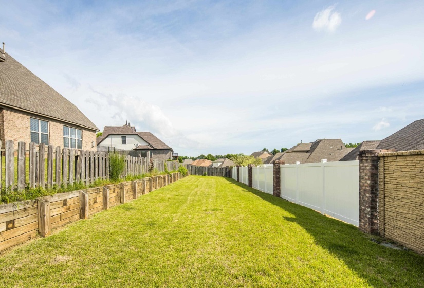 Expansive grassy backyard featuring a wooden retaining wall, a multi-panel wooden fence, and a white vinyl fence with brick pillars