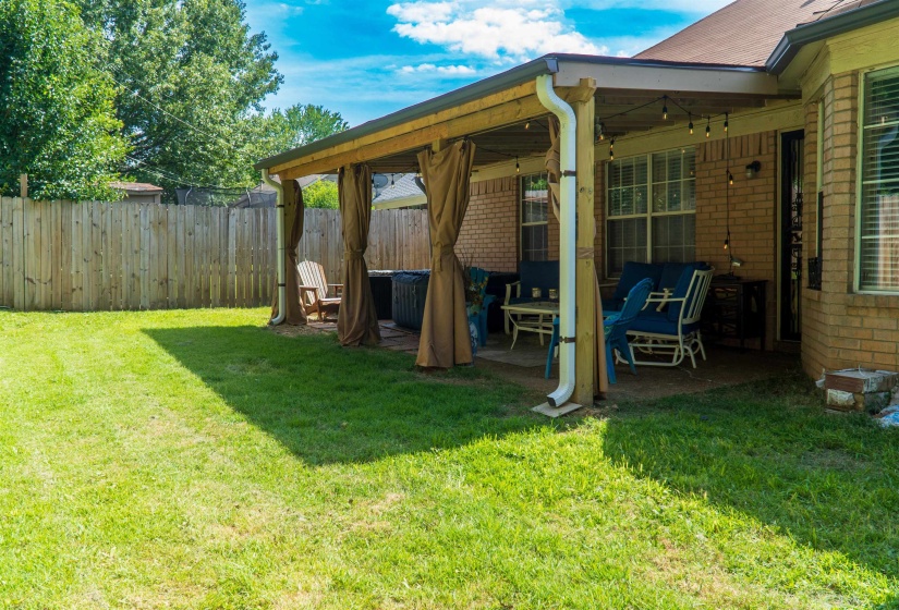 Expansive backyard featuring a covered patio with string lighting, privacy curtains, brick exterior, and wood fencing