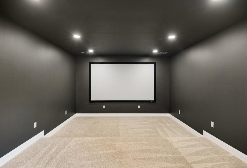 Dedicated media room featuring a large projection screen, dark gray walls, recessed lighting, white baseboards, and neutral-toned carpet flooring