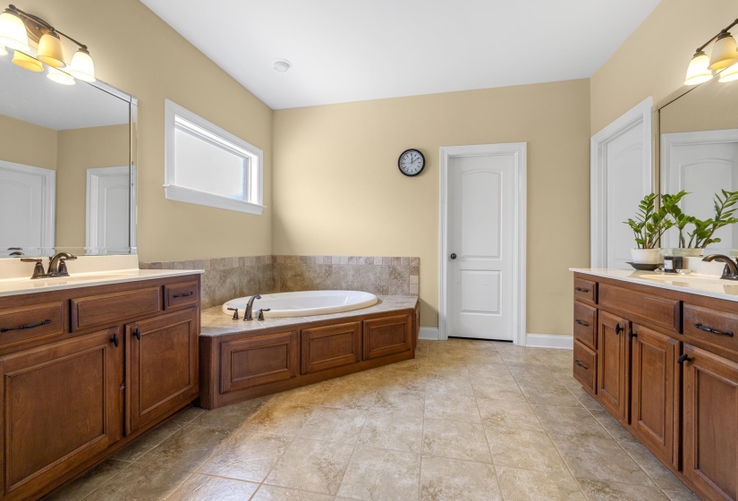 Spacious bathroom featuring a built-in soaking tub with surrounding tile, two individual vanity areas with white countertops and dark wood cabinetry, and neutral-toned floor tiling