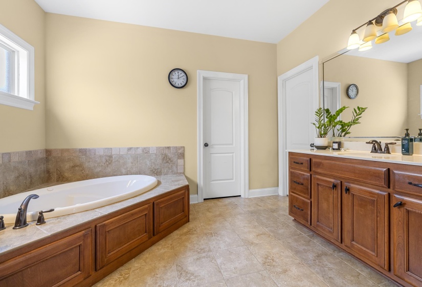 Bathroom featuring a built-in soaking tub with a tiled surround, a window with white trim, and a double vanity with a light-colored countertop and dark wood cabinetry