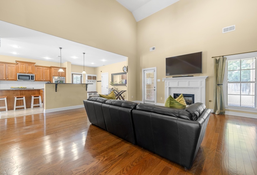 Open-concept living area with vaulted ceilings, wood-finish flooring, a white mantel fireplace, and a kitchen featuring wood cabinetry, stainless appliances, and pendant lighting