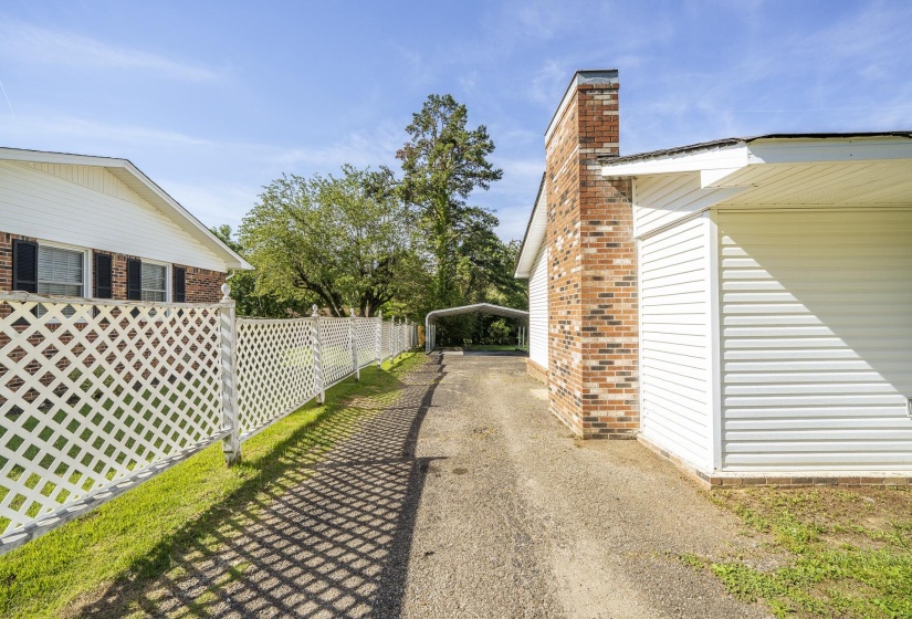 Brick chimney and white siding exterior featuring a paved driveway, a lattice fence, and a distant carport