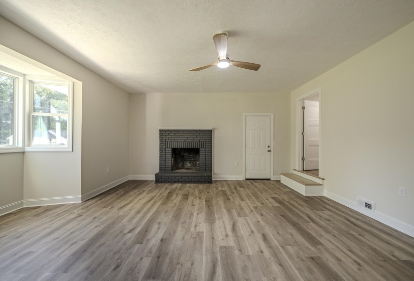 Living area featuring wood-finish flooring, a painted brick fireplace, a bay window, and a ceiling fan with integrated lighting