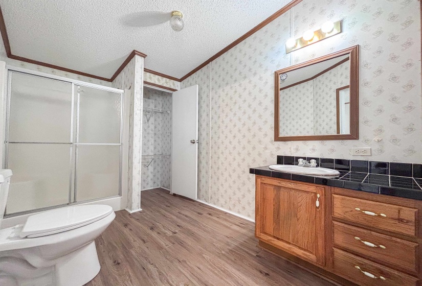 Bathroom featuring wood-finish flooring, a wood vanity with a drop-in sink, a black tiled countertop, a framed wall mirror, and a glass-enclosed shower
