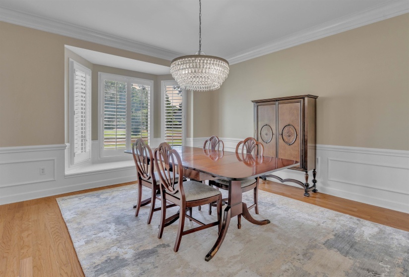 Bright interior featuring a bay window with plantation shutters, a tiered crystal chandelier, wood-finish flooring, crown molding, and wainscoting