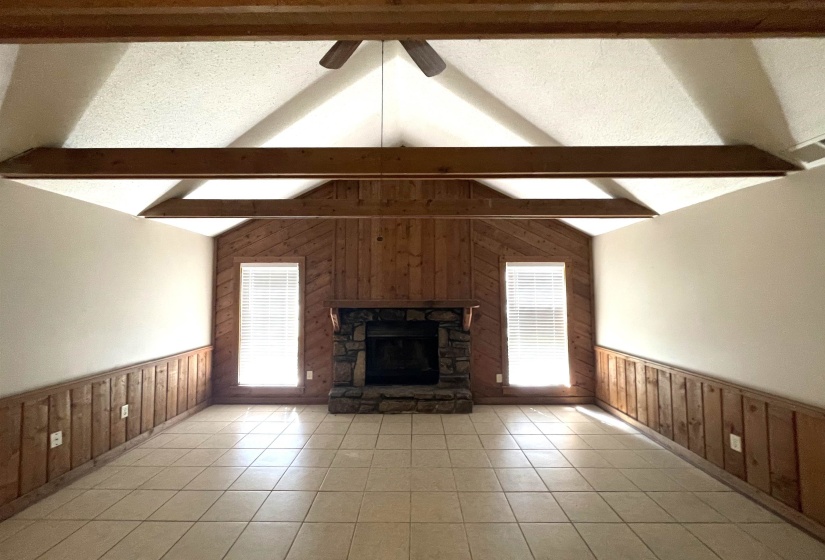 Vaulted ceiling with exposed wood beams, stone fireplace, wood-paneled accent wall, lower wood paneling, and tiled flooring