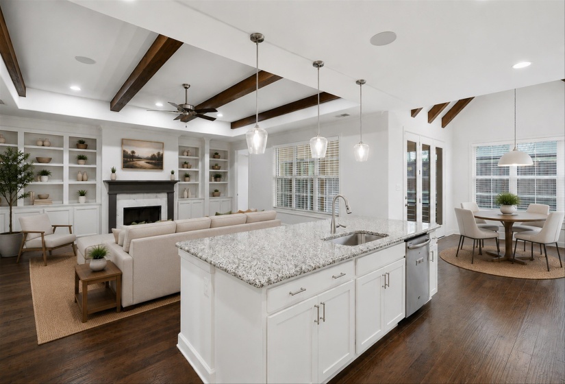 Spacious open-concept living area featuring dark wood-finish flooring, a kitchen island with stone countertop, built-in shelving, exposed ceiling beams, and a fireplace with a dark mantel