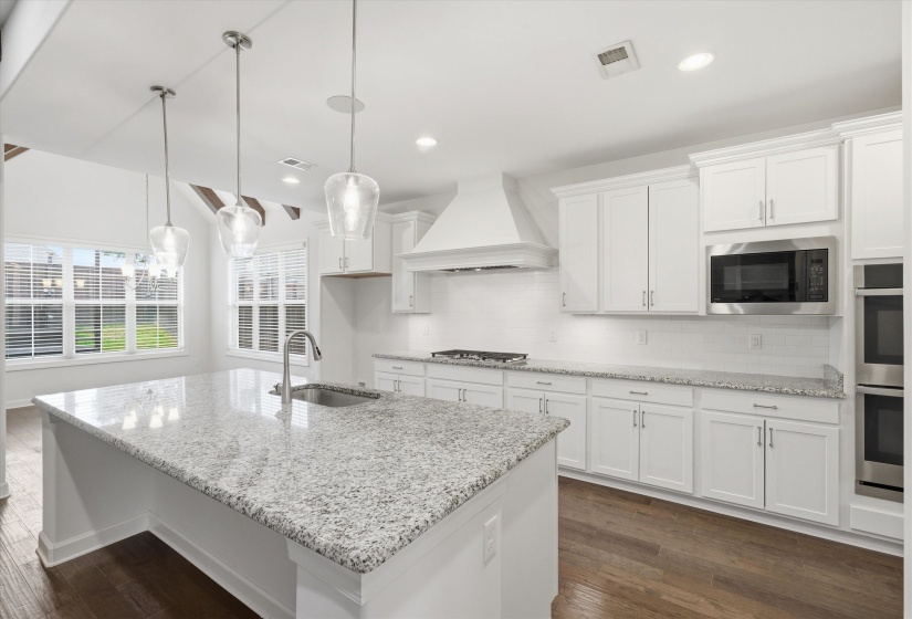 Spacious kitchen featuring an expansive island with a stainless steel undermount sink and three pendant lights, white cabinetry, integrated stainless steel appliances, wood-finish flooring, and multiple windows