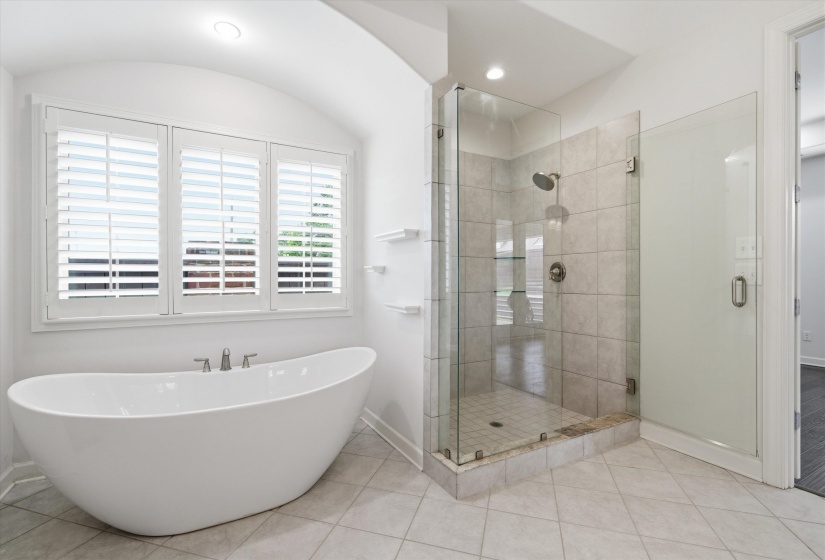 Modern bathroom featuring a freestanding soaking tub, three panel window with plantation shutters, glass-enclosed walk-in shower with tile surround, and light-toned tile flooring