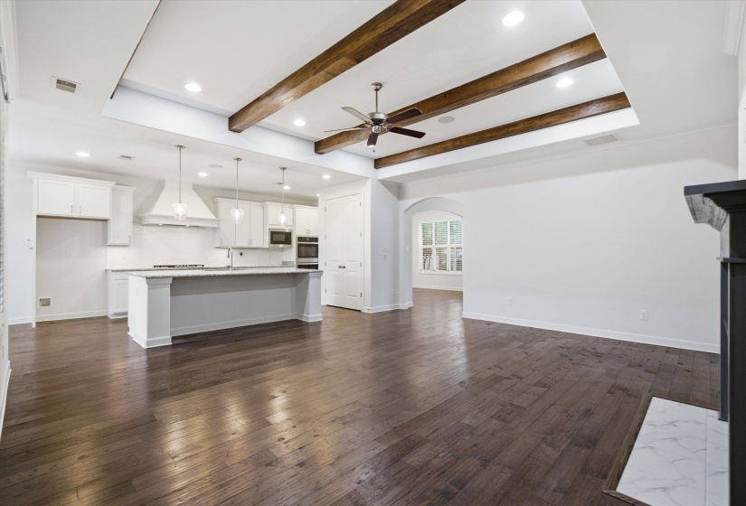 Open-concept living space featuring wood-finish flooring, a kitchen island with pendant lighting, a tray ceiling with exposed wood beams, recessed lighting, and a white tile fireplace hearth