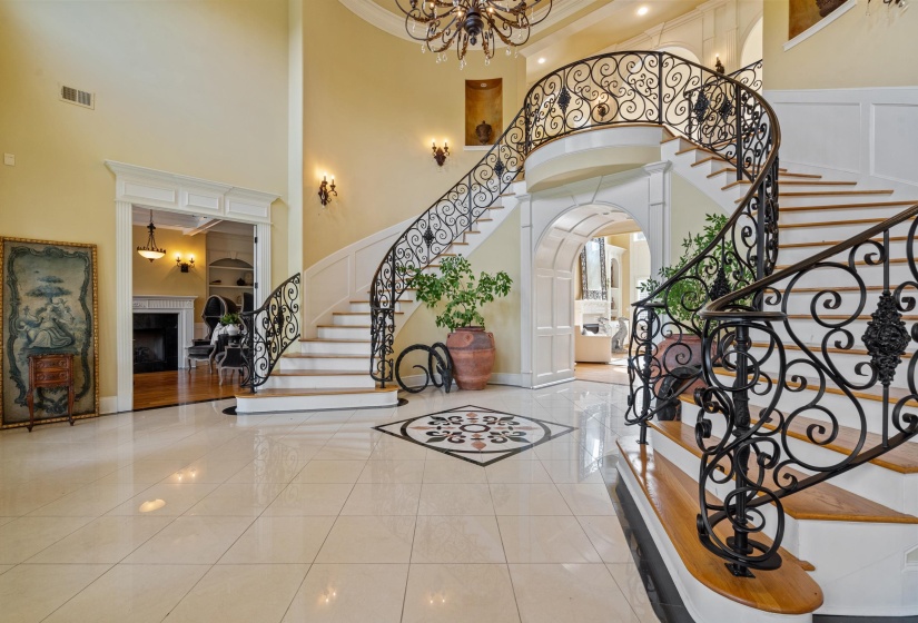 Grand foyer featuring a dual staircase with ornate wrought iron railings, light tile flooring with a central mosaic inlay, and elevated ceilings with recessed lighting