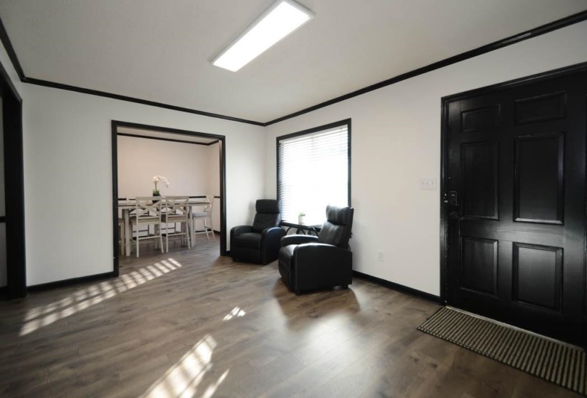Spacious entry area featuring wood-finish flooring, a paneled front door, white walls with dark trim, and a recessed portal to an additional room