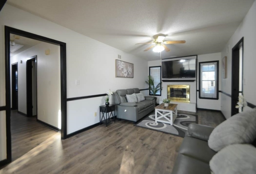 Spacious living area featuring wood-finish flooring, a ceiling fan with integrated lighting, and a prominent fireplace with a gold-toned insert