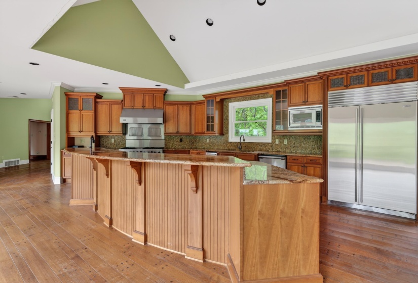 Expansive kitchen featuring a large curved island with wood paneling, wood-finish flooring, and a vaulted ceiling with recessed lighting