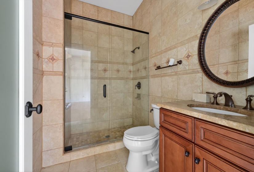 Tiled bathroom featuring a glass-enclosed shower, a vanity with a built-in sink, a circular mirror, and bronze fixtures