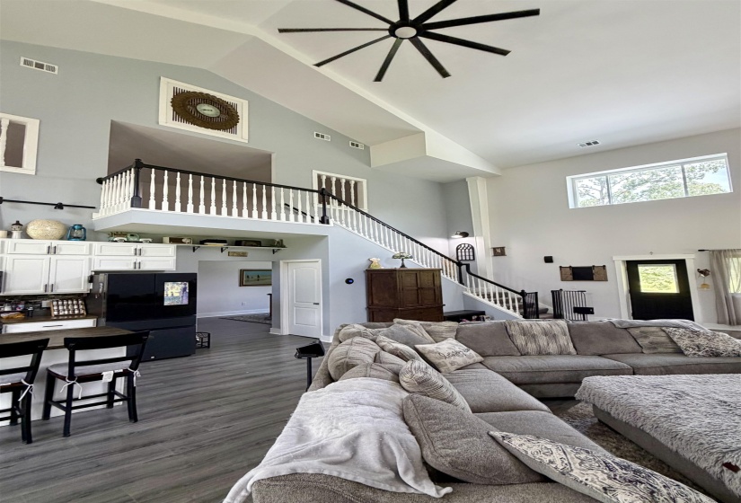 Two-story great room featuring vaulted ceilings, a prominent ceiling fan, a second-story balcony with white balusters and black handrails, and wood-finish flooring