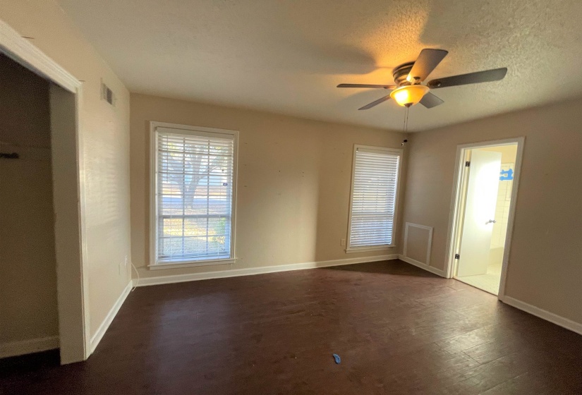 Spacious room featuring dark wood-finish flooring, two windows with blinds, a ceiling fan with integrated lighting, and an open doorway revealing a tiled bathroom with a shower
