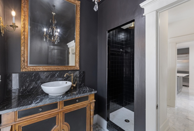 Bathroom featuring a dark tiled shower with white hexagonal floor tiles, a vanity with a black veined countertop and white vessel sink, an ornate gold-framed mirror, and a brass wall sconce