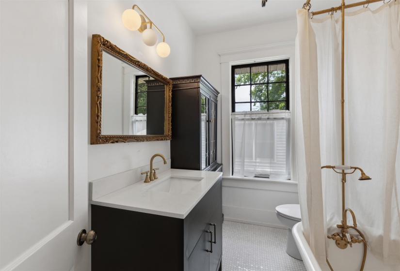 Bathroom featuring a white hex tile floor, a dark wood vanity with a white countertop and gold-finish faucet, a decorative gold-framed mirror, a three-light vanity fixture, and a clawfoot tub with a gold-finish shower fixture
