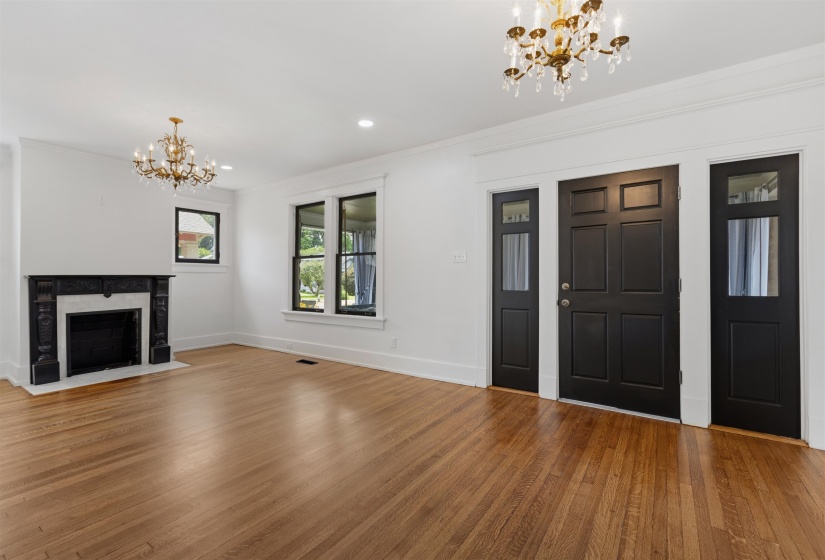 Spacious room featuring hardwood flooring, a decorative fireplace with a black mantel, recessed lighting, and two ornate chandeliers
