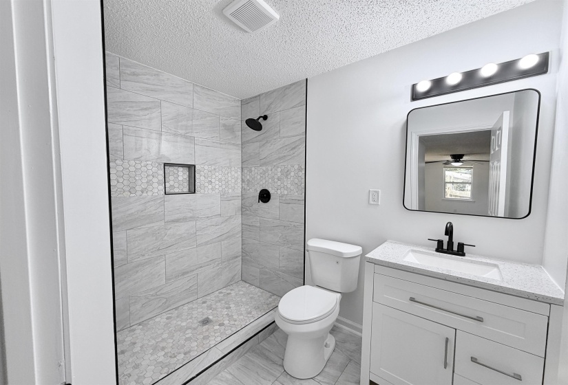 Modern bathroom featuring a walk-in shower with tiled walls and a hex-tile floor, black-finish fixtures, a white vanity with a light-colored countertop, and a contemporary frameless mirror