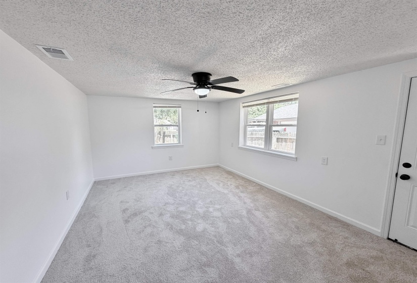Spacious interior room featuring light-colored carpeting, two windows, a ceiling fan with integrated lighting, and a white paneled door with black hardware