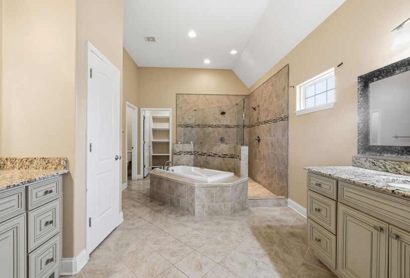 Spacious bathroom featuring a built-in corner soaking tub, a separate tiled shower with glass enclosure, dual vanity sinks with granite countertops, and tiled flooring